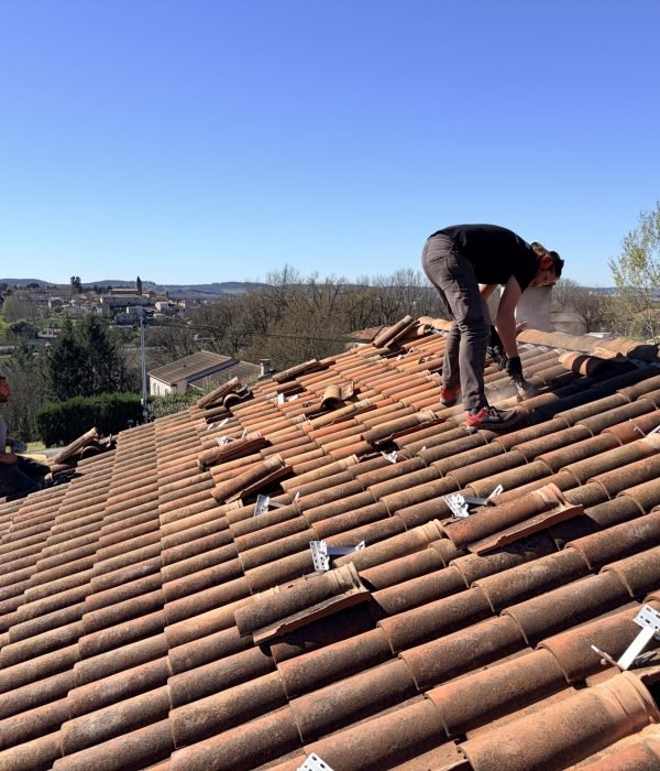 Deux installateurs professionnels d'énergie solaire travaillent sur un grand toit de maison individuelle en tuiles de terre cuite. Au premier plan, un ouvrier accroupi installe des crochets de fixation métalliques. Un deuxième ouvrier est assis plus loin près d'une camionnette de travail. Le ciel est bleu et clair, avec une vue sur un village et une campagne vallonnée en arrière-plan.